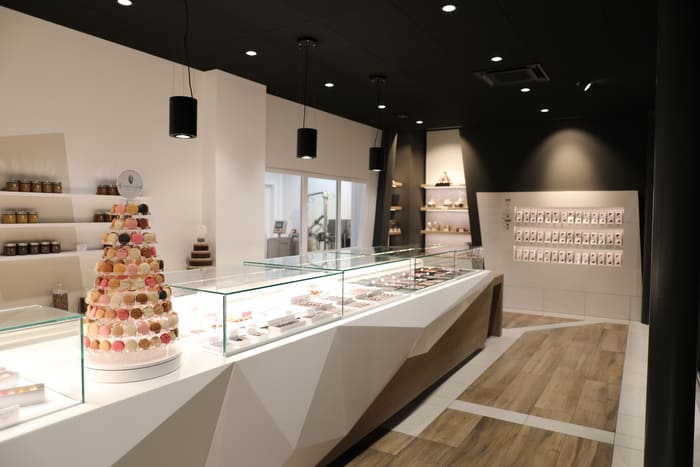 La photo montre une boutique moderne avec un design intérieur épuré, exposant une pyramide de macarons colorés et des présentoirs vitrés contenant des pâtisseries et chocolats.