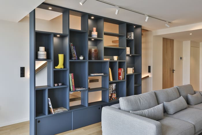 Cet intérieur moderne présente un grand canapé gris devant une bibliothèque sur mesure aux casiers bleu foncé et bois, avec des objets décoratifs et des livres rangés de manière esthétique.