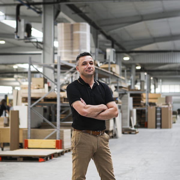 Un homme en polo noir et pantalon beige se tient les bras croisés dans un entrepôt rempli de boîtes et de matériel d'expédition, affichant un sourire confiant.